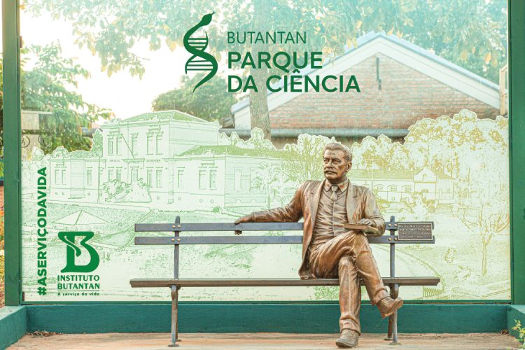 Estátua do cientista Vital Brazil sentado em banco de madeira com fundo do Parque da Ciência Butantan